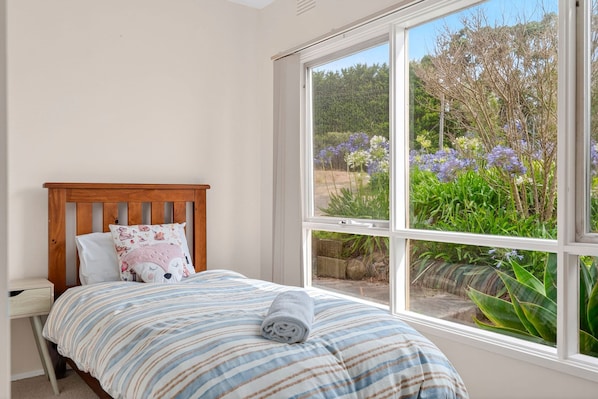 A bedroom with a wooden headboard, a bed with striped bedding, a pillow with a dog design, and a view of a garden through a large window.