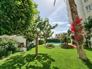 A well-maintained garden with a swimming pool, palm trees, and a building in the background. - Cozy apartment with terrace and pool in L'Escala (L'Escala)