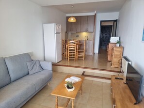 A compact living space with a sofa, a wooden table with a plant, a TV, and a kitchenette in the background.