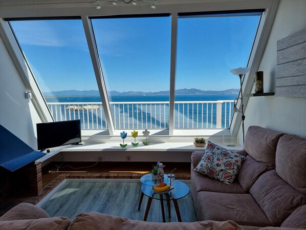 A balcony with a view of the sea and mountains, a sofa, a small table with fruit, and a TV.