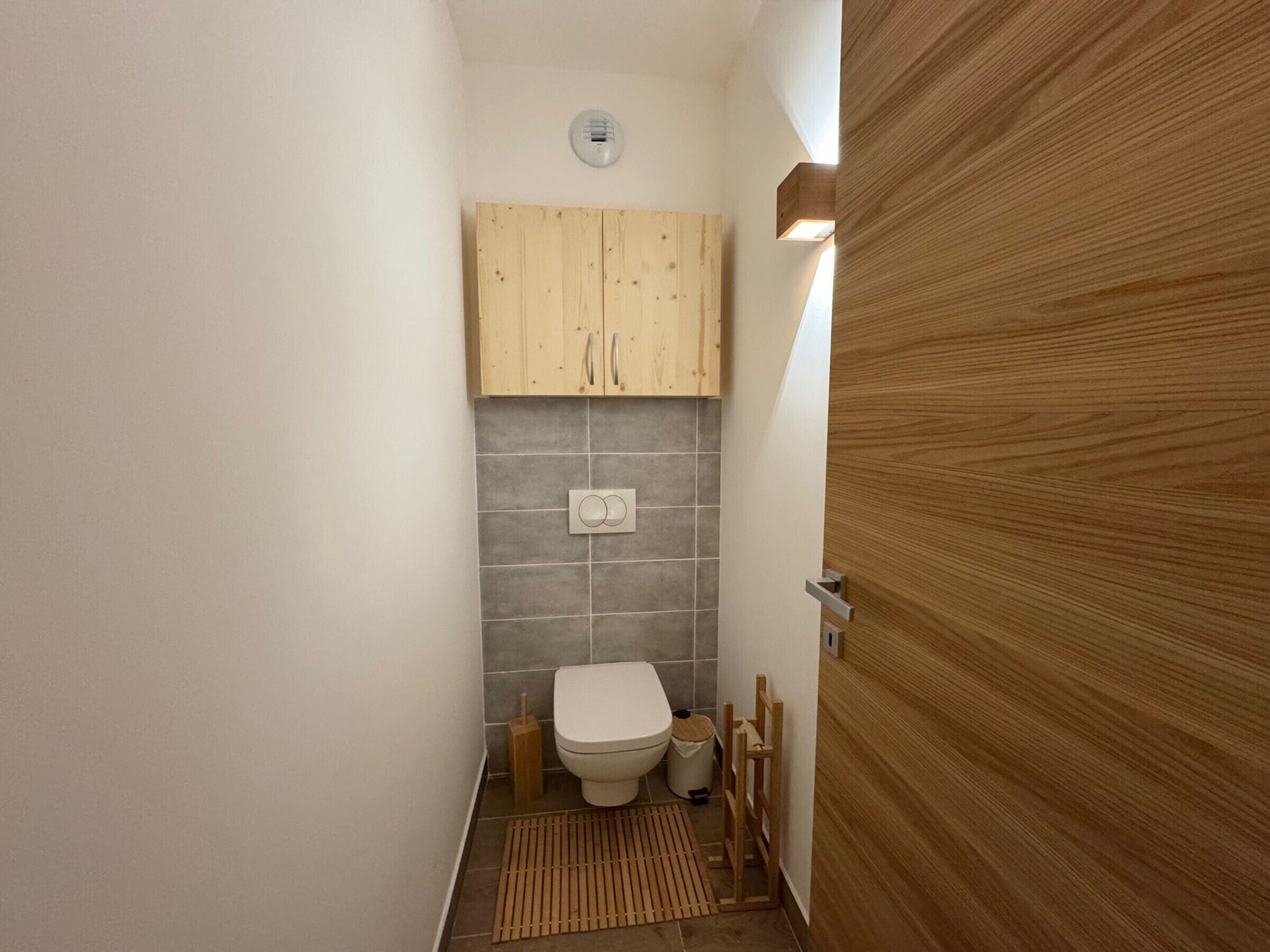 A small bathroom with a toilet, a wooden cabinet above the sink, and a wooden door.