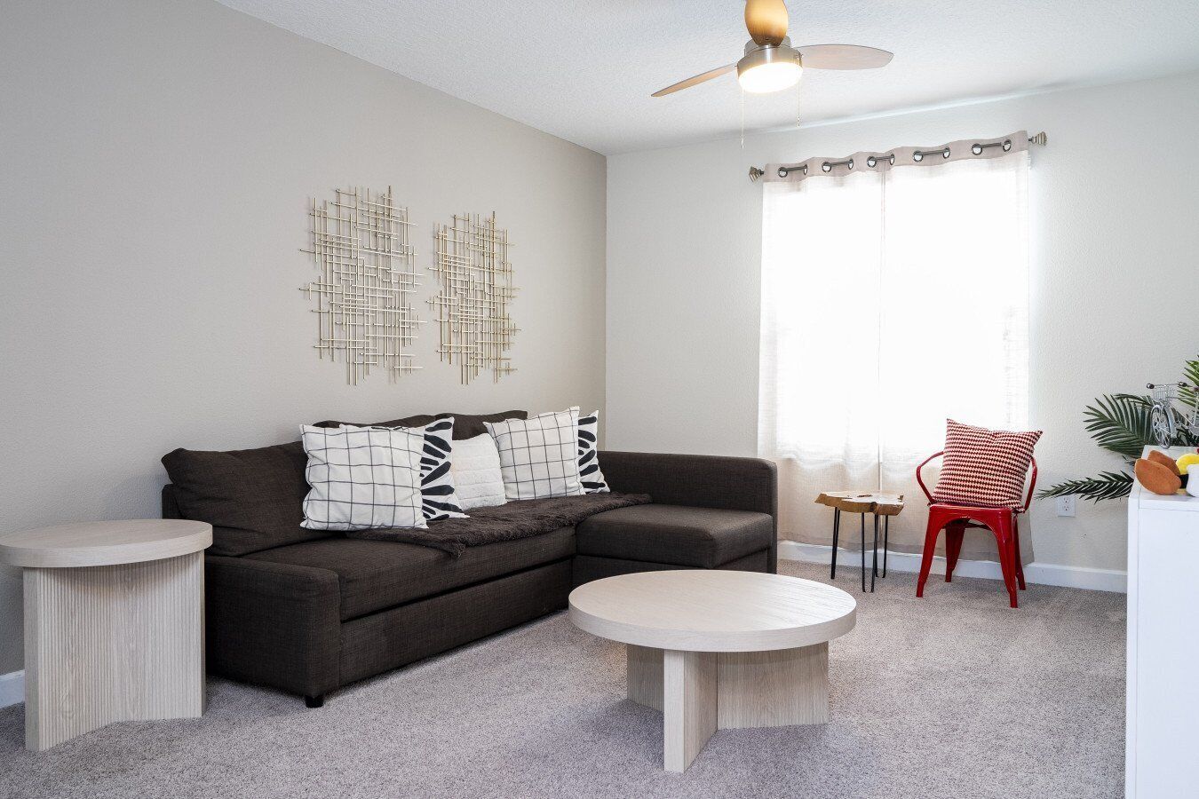 Ein modernes Wohnzimmer mit einem Ecksofa, zwei Beistelltischen, einem runden Couchtisch, einem Deckenventilator und einem Fenster mit Vorhängen.
