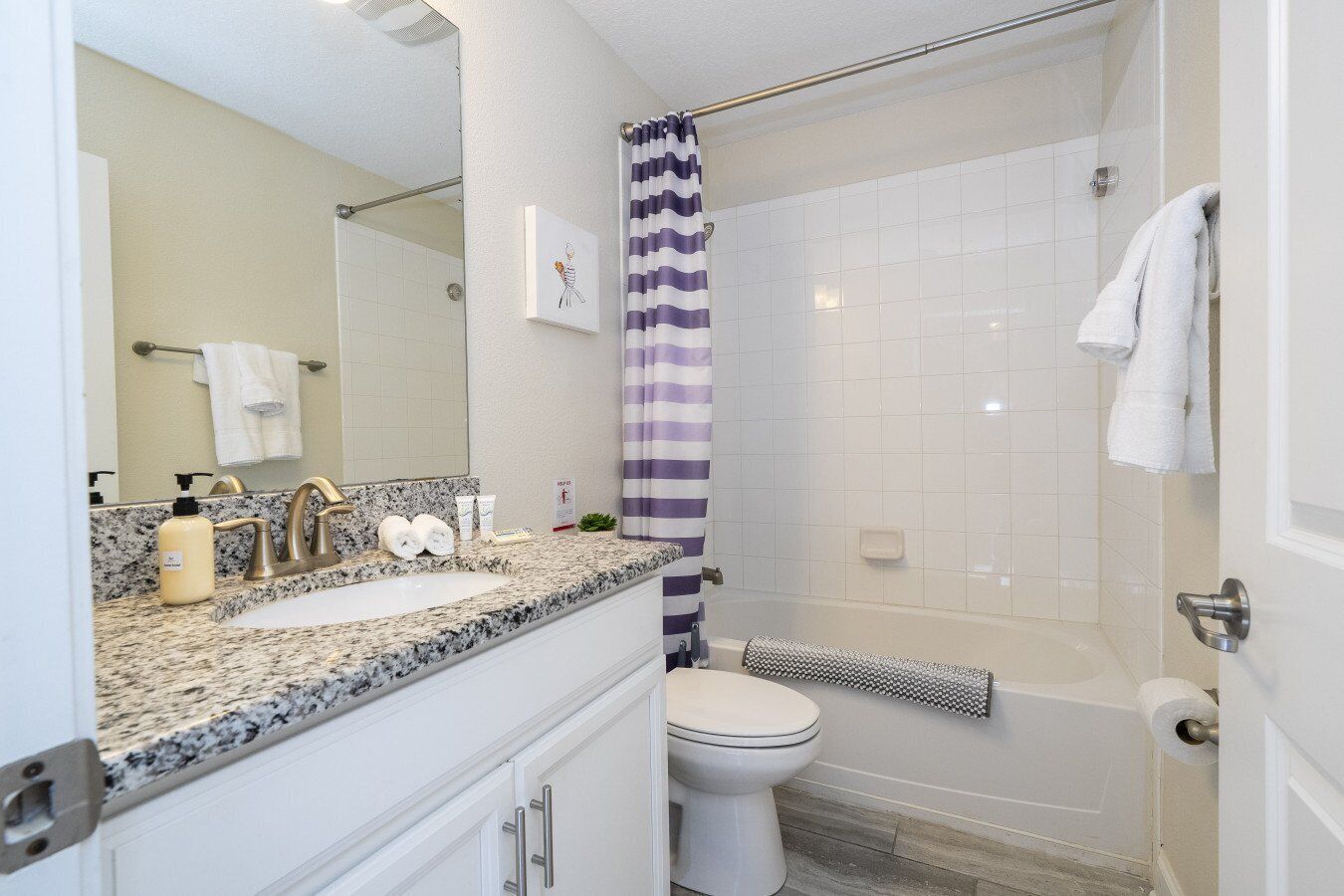 A bathroom with a granite countertop sink, a white toilet, a shower with a purple and white striped curtain, and a towel rack with white towels.