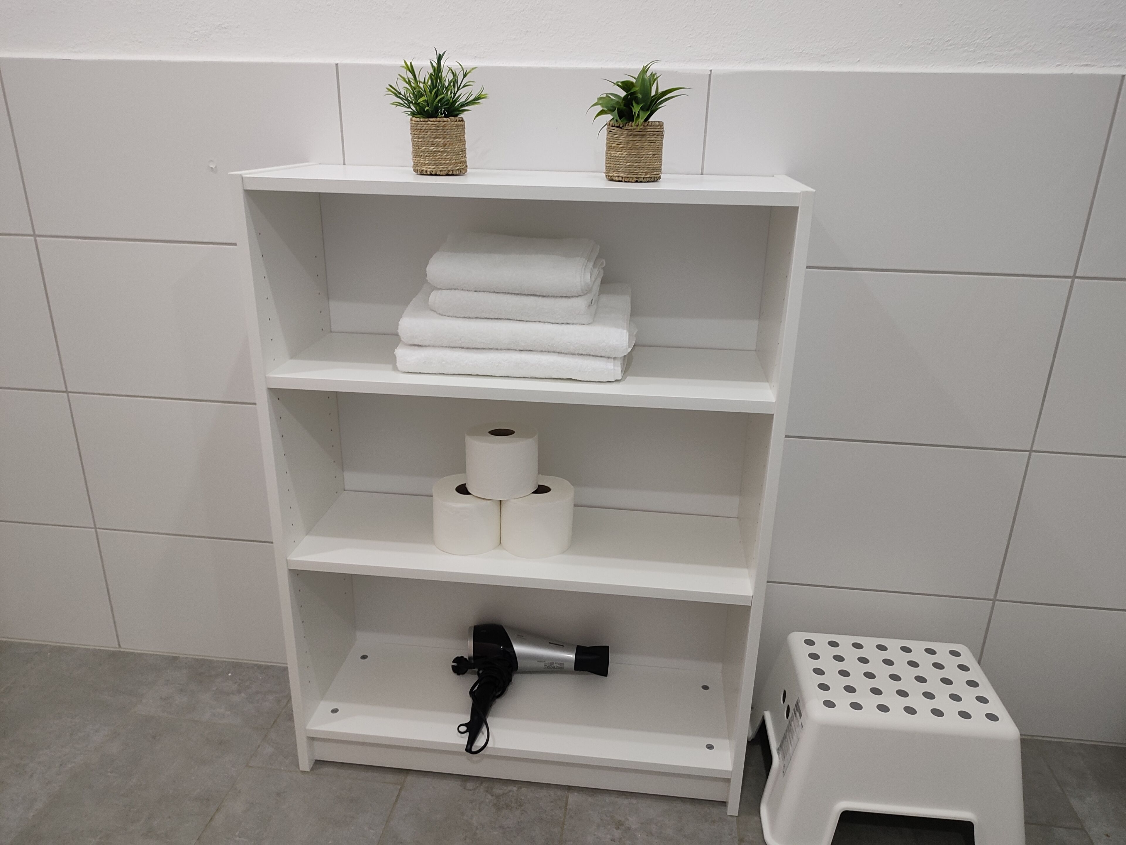 Une étagère blanche contenant des serviettes, du papier toilette, un sèche-cheveux et un panier à linge.