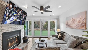 A modern living room with a fireplace, a ceiling fan, a flat-screen TV, a large window, and a painting on the wall. - Walk to PCMR • Chic Top-Floor Mountain Condo (Park City)