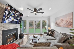Ein modernes Wohnzimmer mit Kamin, Deckenventilator, Flachbildfernseher, großem Fenster und einem Gemälde an der Wand.