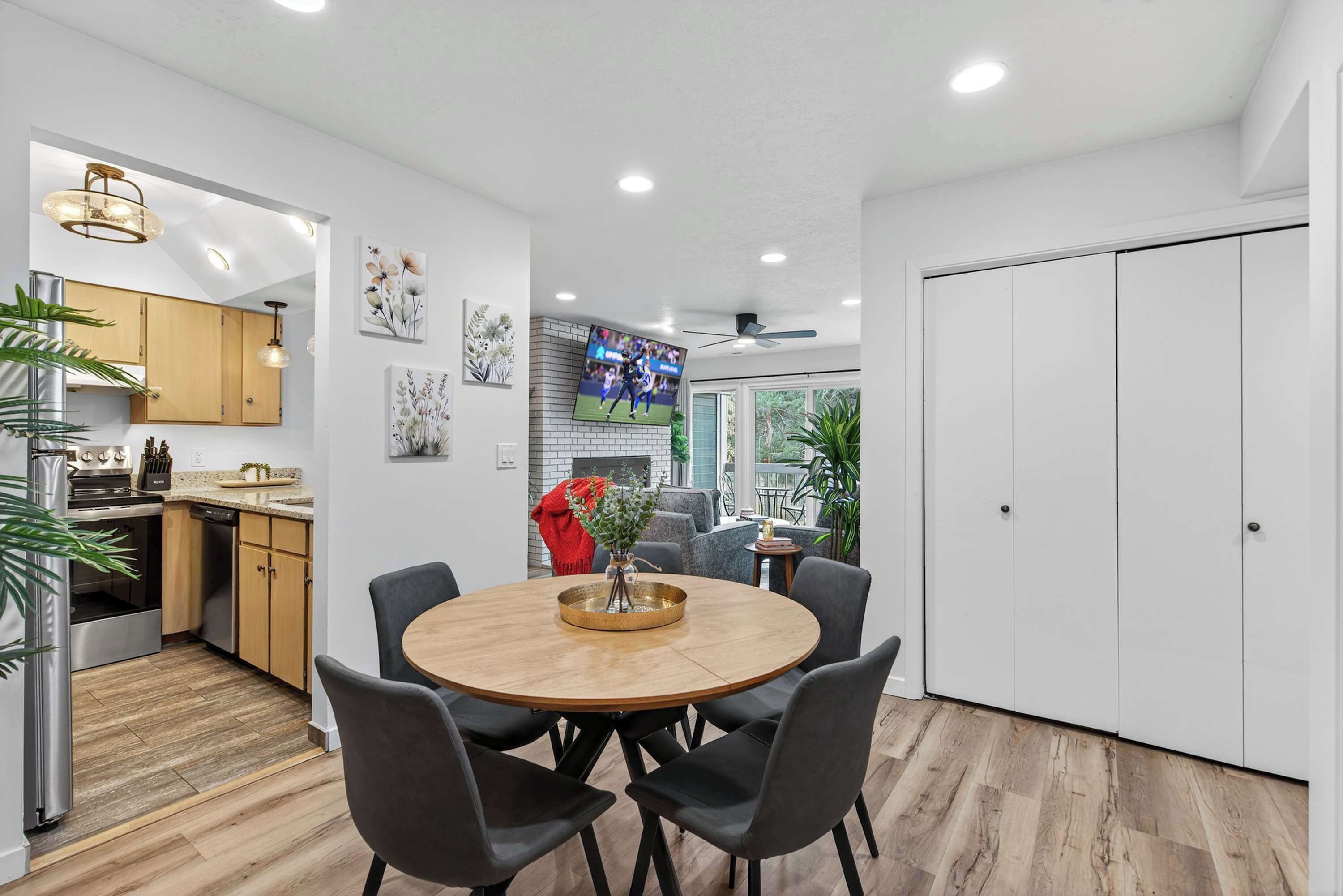A modern kitchen with wooden cabinets, a dining area with a round table and chairs, and a TV mounted on the wall.