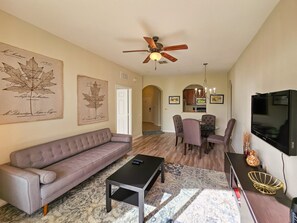 A living room with a sofa, coffee table, TV, and dining area with chairs and a chandelier. - Last Minute Disney Getaway! Luxury Condo with Private Balcony. Book Now! #114235 (Orlando)