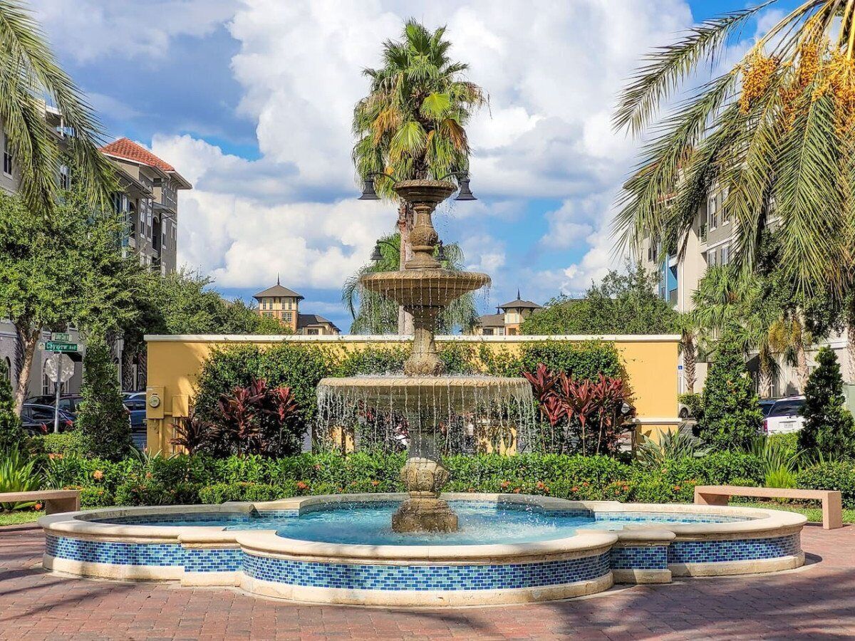 A fountain with multiple tiers and a pool in the foreground, surrounded by lush greenery and buildings in the background.