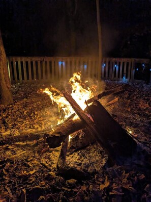 A campfire with logs and burning wood, surrounded by dry leaves on the ground. - Wooden Oasis minutes from Burdette & USI (Evansville)