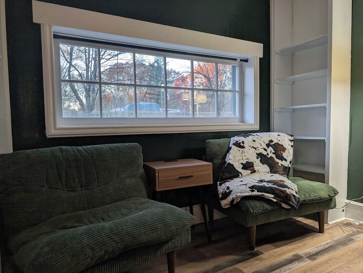 A room with a large window, a green sofa, a chair with a cowhide throw, and a small wooden side table.