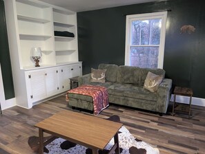 A living room with a sofa, ottoman, and coffee table, a white cabinet with shelves, and a window with a view. - Wooden Oasis minutes from Burdette & USI (Evansville)