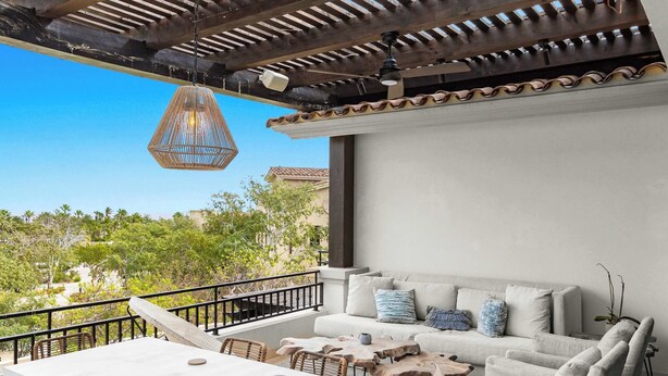 Un balcon avec une table blanche et des chaises en osier, un canapé avec des coussins et un luminaire suspendu.