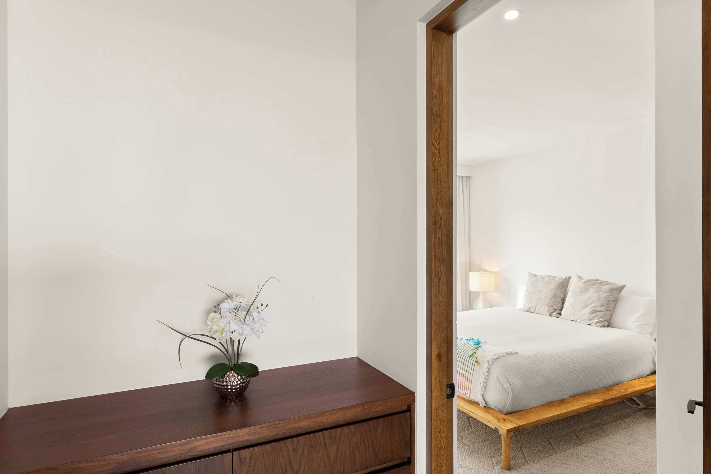 A bedroom with a wooden bed frame, a bedside table with a vase of flowers, and a large mirror reflecting the room.