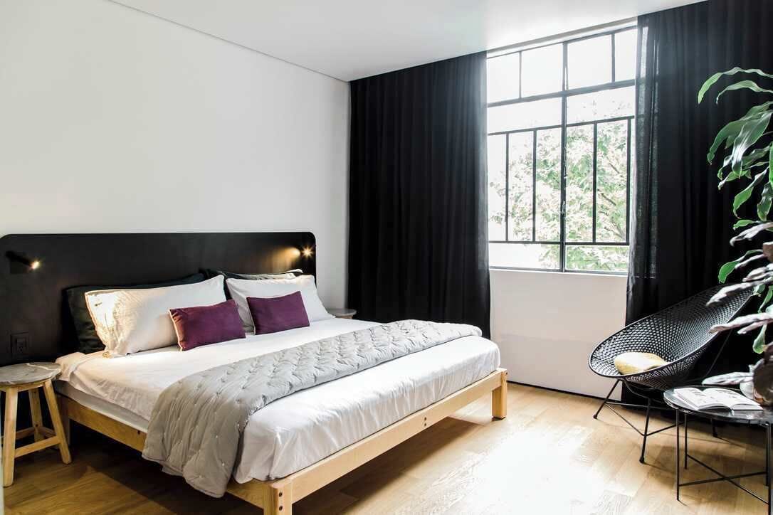 Un dormitorio moderno con una cama de madera, un taburete pequeño, una silla de metal negro y un amplio ventanal con cortinas negras.