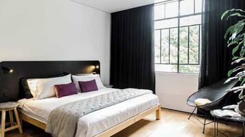 Un dormitorio moderno con una cama de madera, un taburete pequeño, una silla de metal negro y un amplio ventanal con cortinas negras.