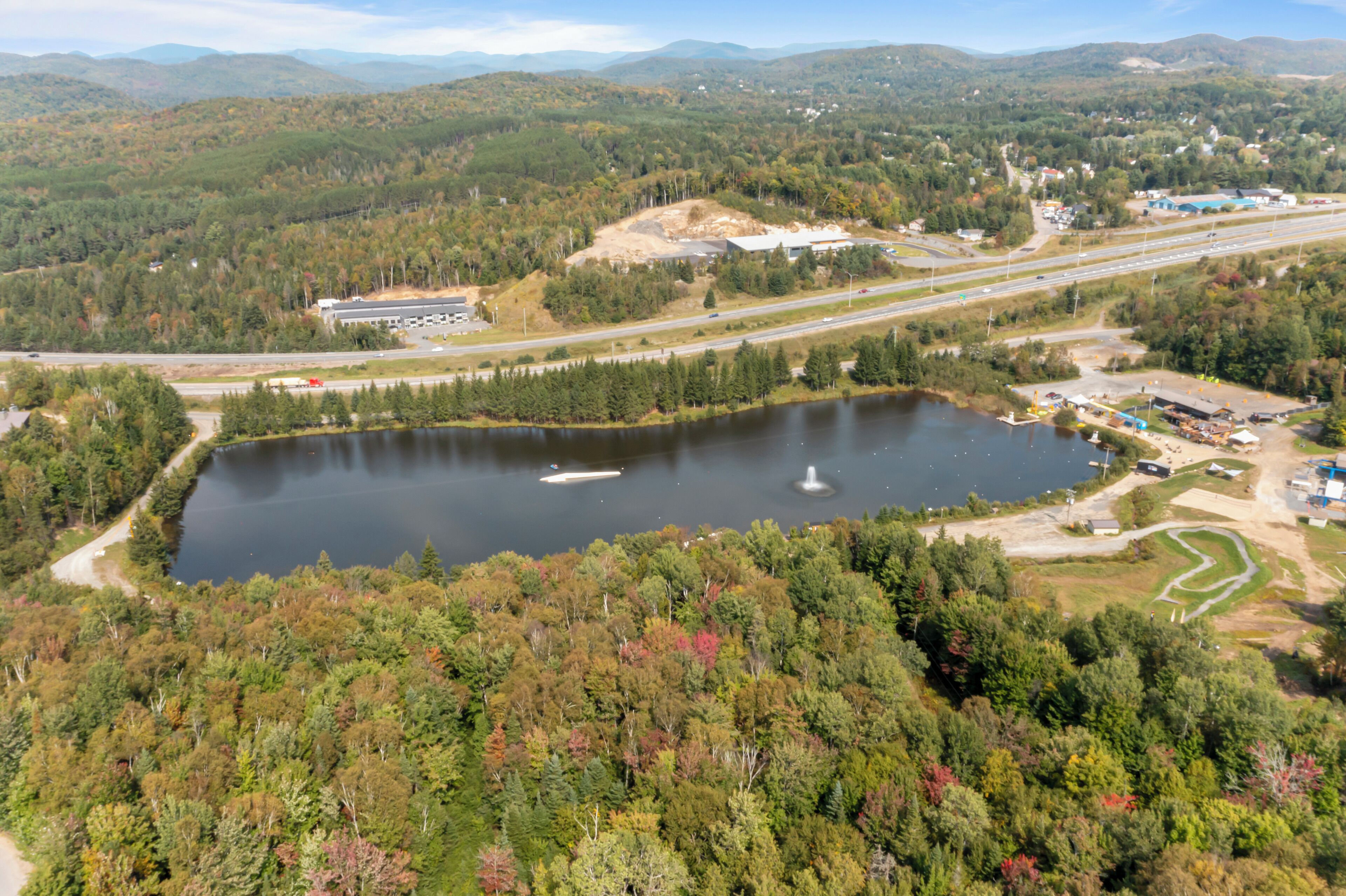 An aerial view of a lake surrounded by dense forest, with a highway and industrial buildings in the background.