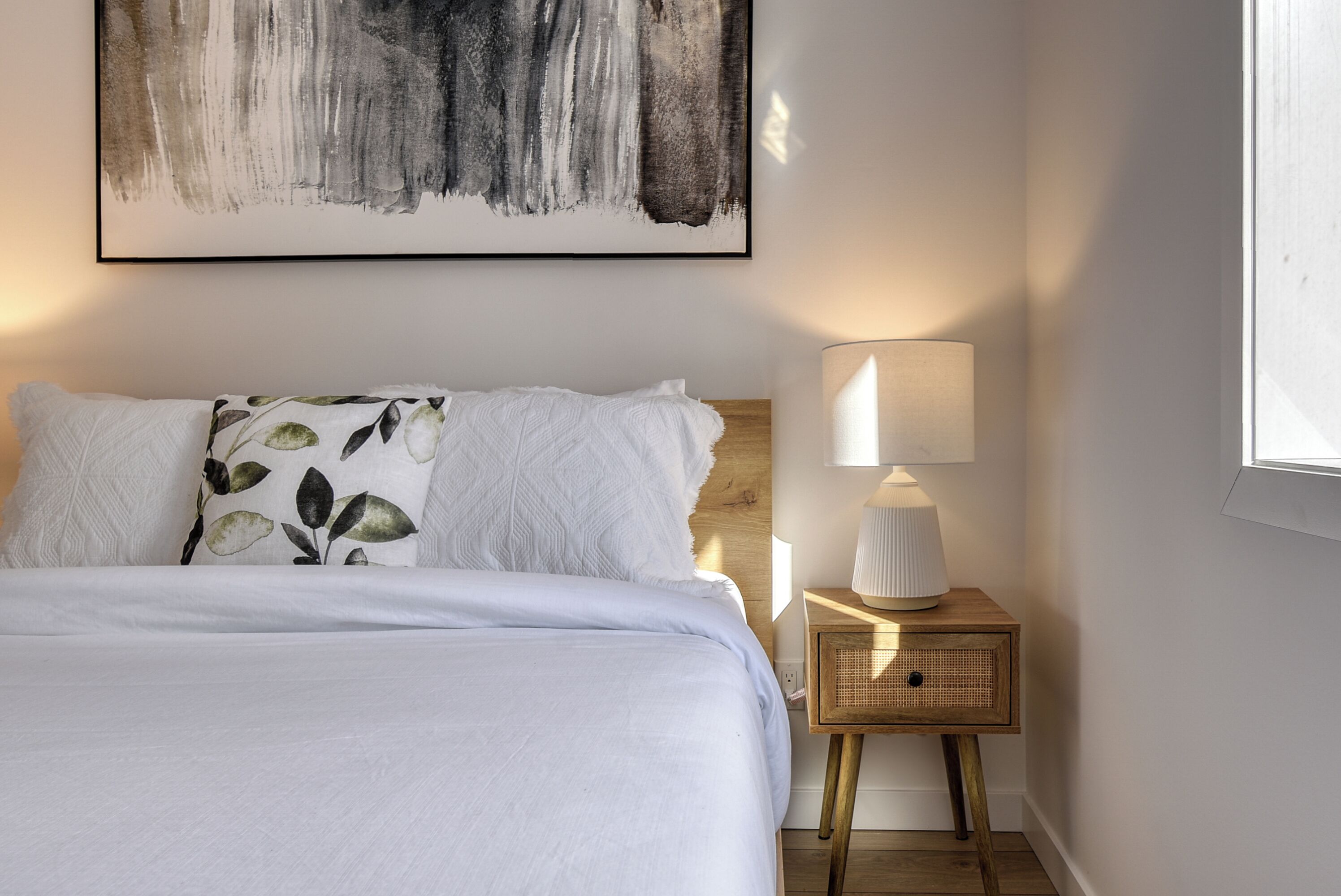 Un lit bien fait, avec du linge de lit blanc et un coussin orné d’un motif de feuilles, une lampe de chevet posée sur une table de nuit en bois, et un grand tableau abstrait accroché au mur.