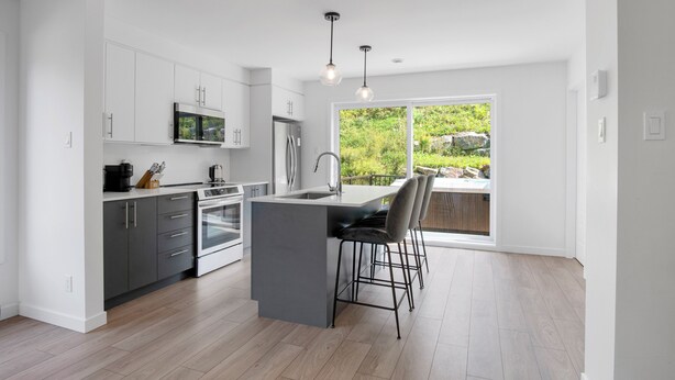 A modern kitchen with white cabinets, a central island, bar stools, and a large window offering a view of greenery. - Le Relais Mont-Blanc (Les Laurentides)