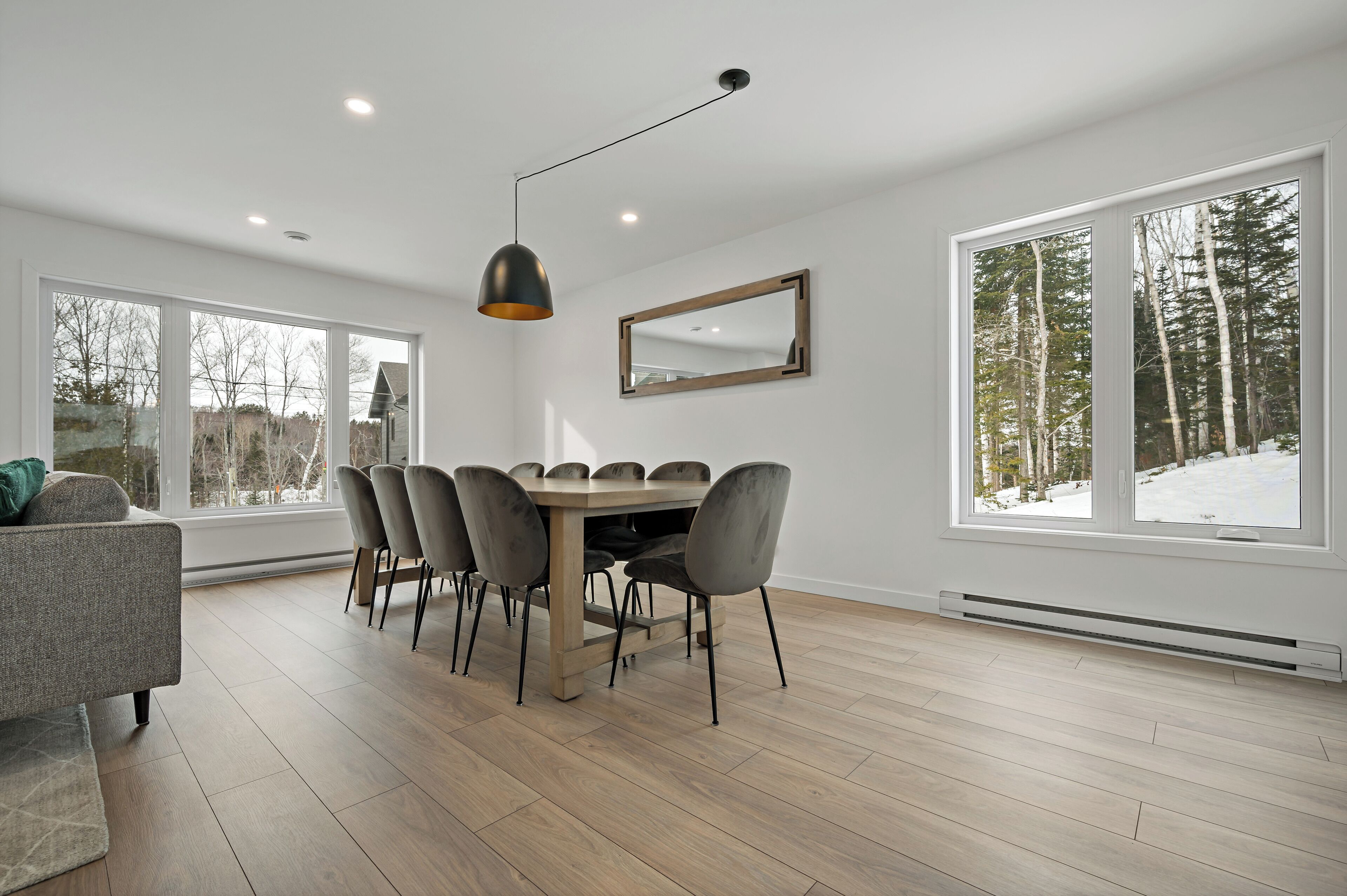 A modern dining area with a wooden table and chairs, large windows with a view of trees, and a ceiling light fixture.