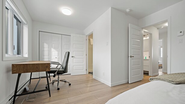Een moderne slaapkamer met een houten bureau, een zwarte bureaustoel en een bed met een witte dekbedovertrek.