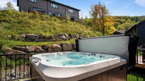 A hot tub on a wooden deck with a view of a hillside and a modern building in the background. - Le Relais Mont-Blanc (Les Laurentides)