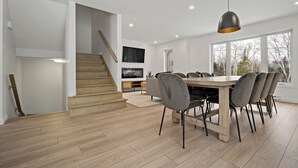 A modern kitchen with a wooden staircase, a dining area with a large table and chairs, and a view through large windows. - Le Relais Mont-Blanc (Les Laurentides)