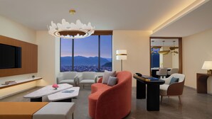 Et moderne hotelværelse med et stort vindue der udover byudsigt, et fladskærms-tv monteret på væggen, og moderne møbler inklusive en rød lænestol og en hvid sofa.