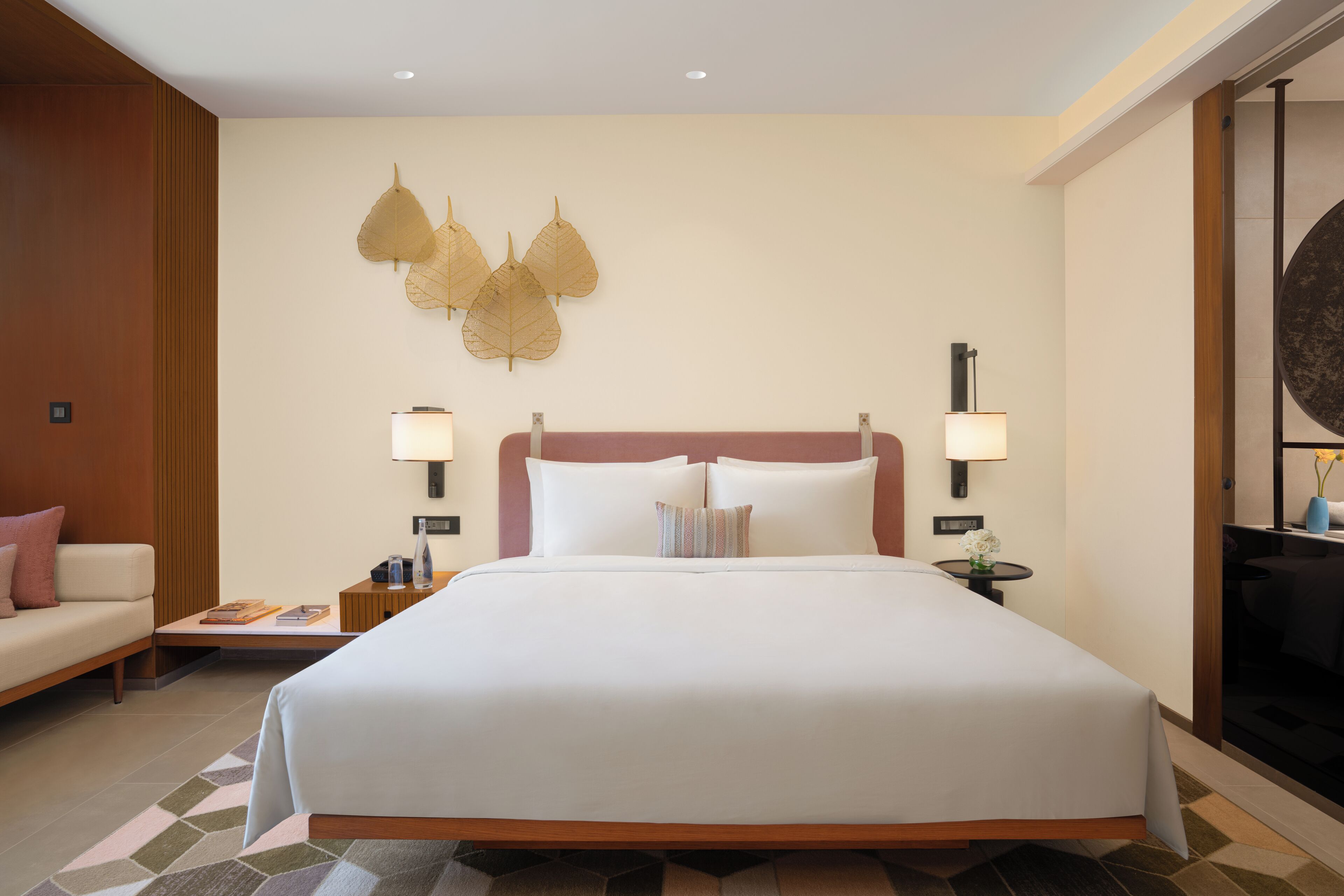 A hotel room with a large bed, two bedside tables, a nightstand, a wall-mounted lamp, and a decorative leaf wall art piece.