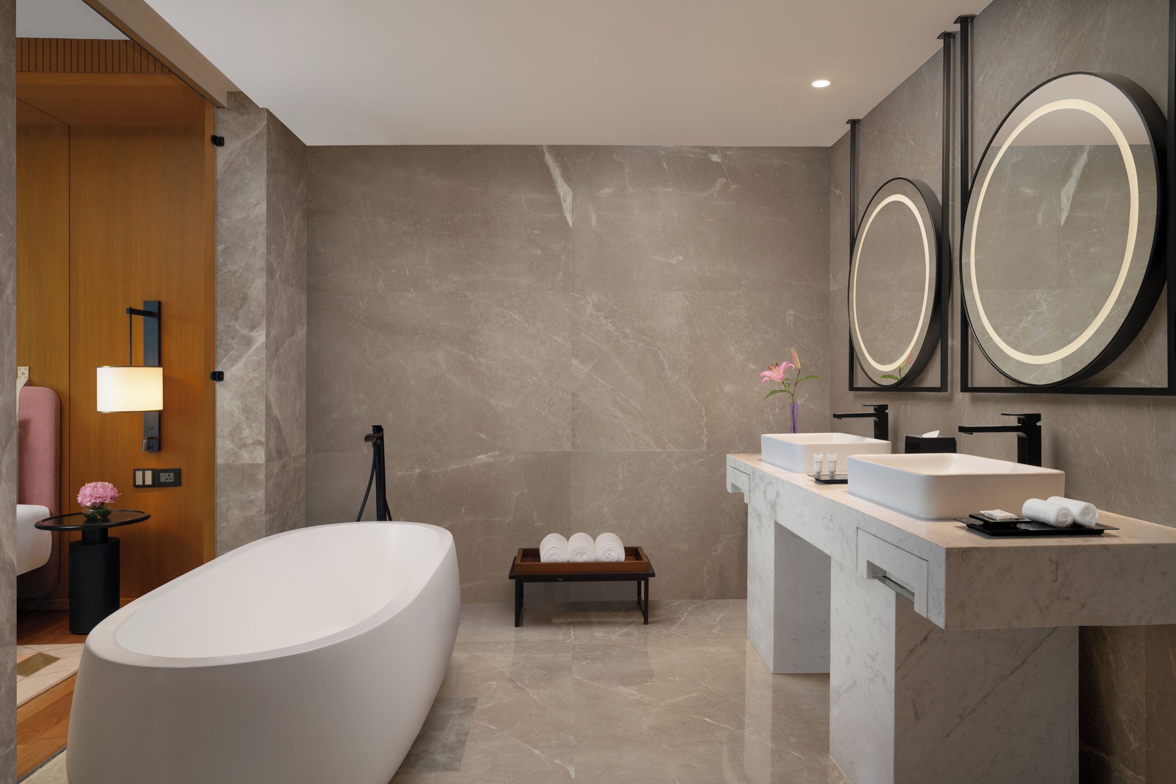 A modern bathroom with a freestanding bathtub, a vanity with a large mirror, and a bench with towels.