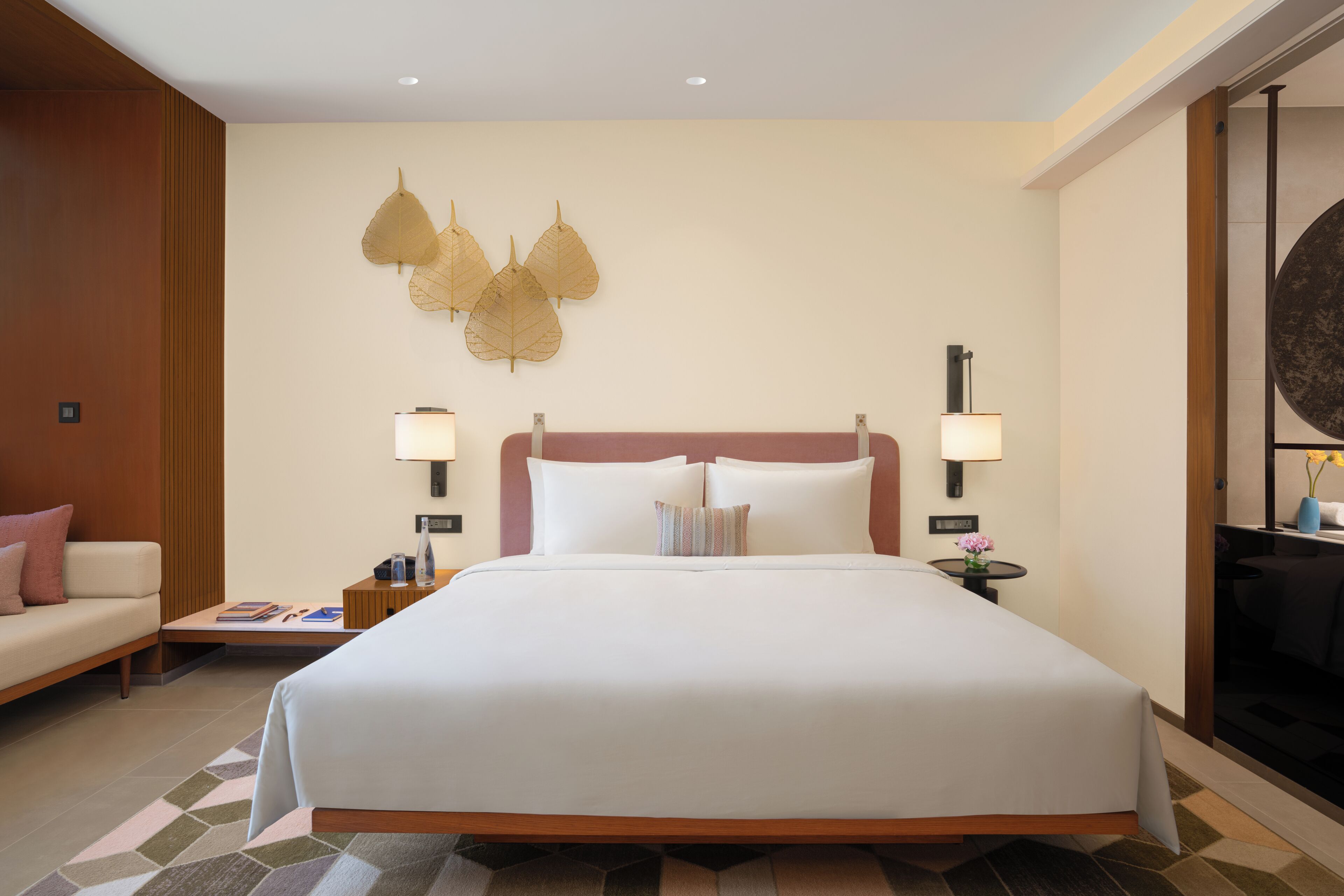 Quarto de hotel com uma cama grande, duas mesas de cabeceira, um criado-mudo, um sofá e um objeto decorativo na parede.
