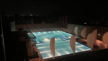 Una piscina en la azotea con cerramiento de cristal, rodeada de elementos arquitectónicos modernos y vista al cielo nocturno.