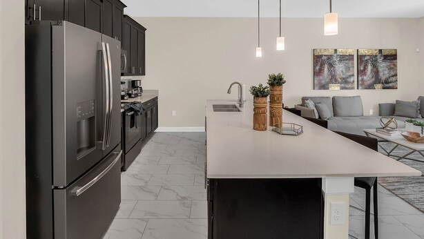 A modern kitchen with dark cabinets, a stainless steel refrigerator, and a marble floor. There is a dining area with a long table and two pendant lights. - Last Minute Disney Getaway! Private Pool & Themed Bedrooms. Book Now! #150581 (Orlando)