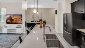 A modern kitchen with a central island, stainless steel appliances, pendant lighting, and a dining area with a large painting. - Last Minute Disney Getaway! Private Pool & Themed Bedrooms. Book Now! #150581 (Orlando)