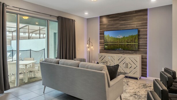 A modern living room with a grey sofa, a flat-screen TV mounted on a wooden wall, a glass sliding door leading to a balcony, and a white cabinet with decorative patterns. - Last Minute Disney Getaway! Private Pool & Themed Bedrooms. Book Now! #150903 (Orlando)