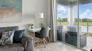 En moderne stue med en grå sofa, et hvidt skrivebord og en grå stol. Der er et stort vindue med udsigt over en park og en bygning i baggrunden.