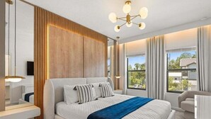 Ein modernes Schlafzimmer mit einem großen Bett, einer Wand in Holzoptik, einer Chandelierleuchtreihe und Blick ins Freie.