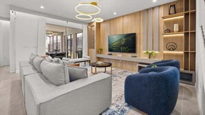 Een moderne woonkamer met een grijze bank, een blauwe fauteuil, een aan de muur bevestigde televisie en een houten wandplank met decoratieve voorwerpen.