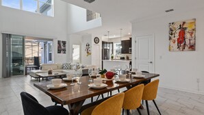 Un'ampia zona pranzo con soffitto alto, una grande finestra e una cucina moderna sullo sfondo.