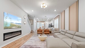 Uma sala de estar moderna com lareira, uma televisão grande e um espaço de jantar com cozinha ao fundo.