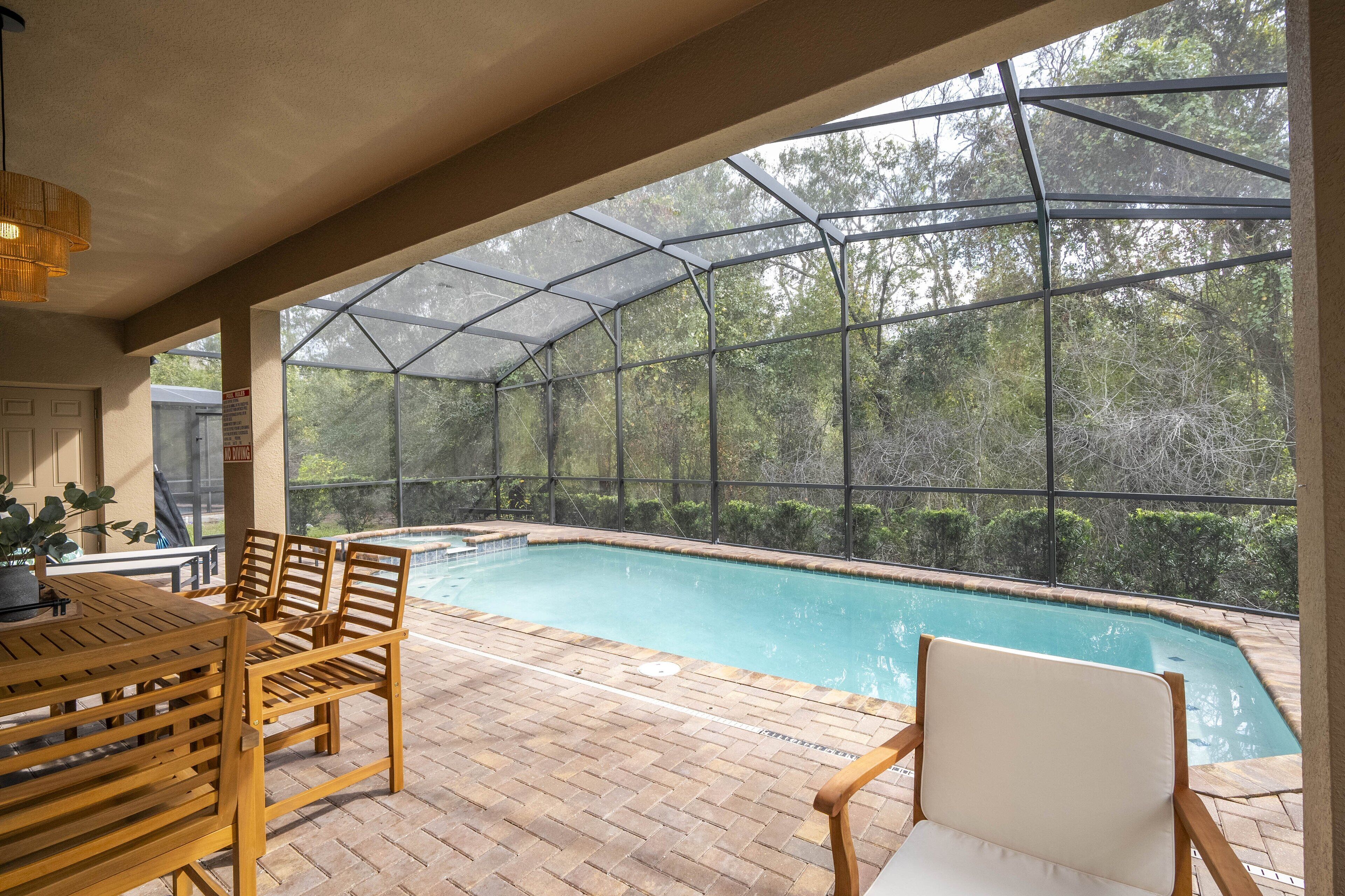 Un espace extérieur abrité avec une piscine, comprenant une table et des chaises en bois, le tout entouré d’une structure en verre.