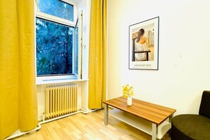 A room with a window, a radiator, a wooden table with a vase, and a framed picture on the wall.
