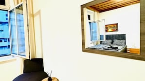 Ett rum med en säng, en stol, ett litet bord, ett fönster och en spegel som reflekterar rummet.