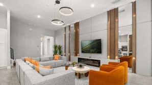 Ein modernes Wohnzimmer mit einem grauen Ecksofa, orangefarbenen Sesseln, einem Flachbildfernseher und einem Glastisch.