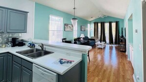 A modern kitchen with a central island, a dining area with a sofa, and a living room with a TV. - Top-Floor River Oaks Escape  2BR, 2BA (Myrtle Beach)