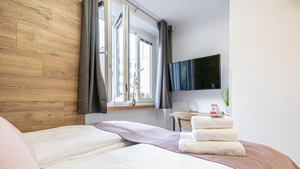 Une chambre d’hôtel avec une tête de lit en bois, un lit avec du linge de maison blanc, un bureau avec une chaise, un téléviseur à écran plat fixé au mur et une fenêtre avec des rideaux.