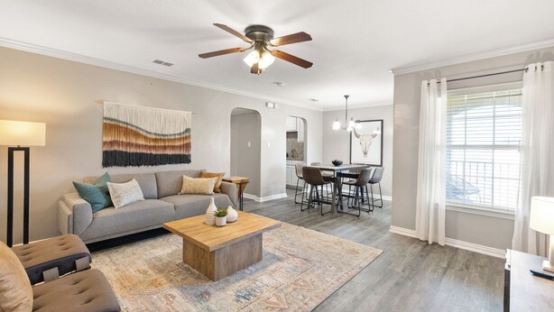 Un salon spacieux avec un canapé gris, une table basse en bois et un tapis à motifs. On y trouve également un coin repas avec une table et des chaises, et un ventilateur de plafond au-dessus.