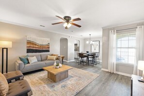 A spacious living room with a grey sofa, a wooden coffee table, and a patterned rug. There is a dining area with a table and chairs, and a ceiling fan above. - Texas Two-Step Retreat Walk to AT&T & Globe Life (Arlington)