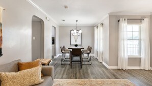 Un salon spacieux avec un canapé gris, un tapis à motifs, un coin repas comprenant une table et des chaises, et une grande fenêtre avec des rideaux.