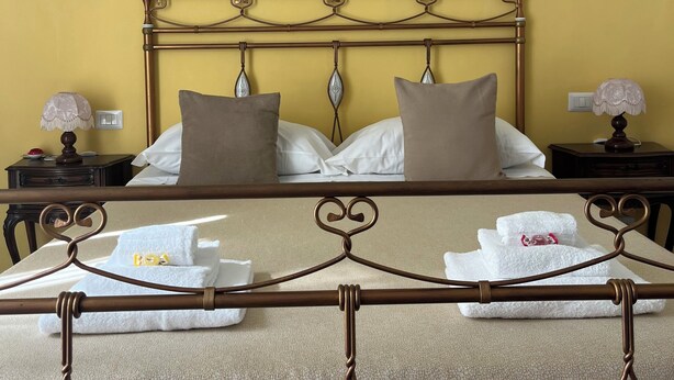 Un letto ben rifatto con una testiera in metallo, cuscini bianchi e una coperta beige. Sono presenti due comodini con lampade e un decoro a parete.